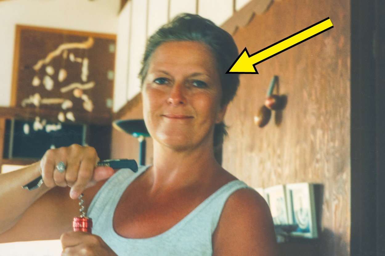 Person smiling while using a corkscrew, highlighted by an arrow. Casual setting with wall decorations in the background