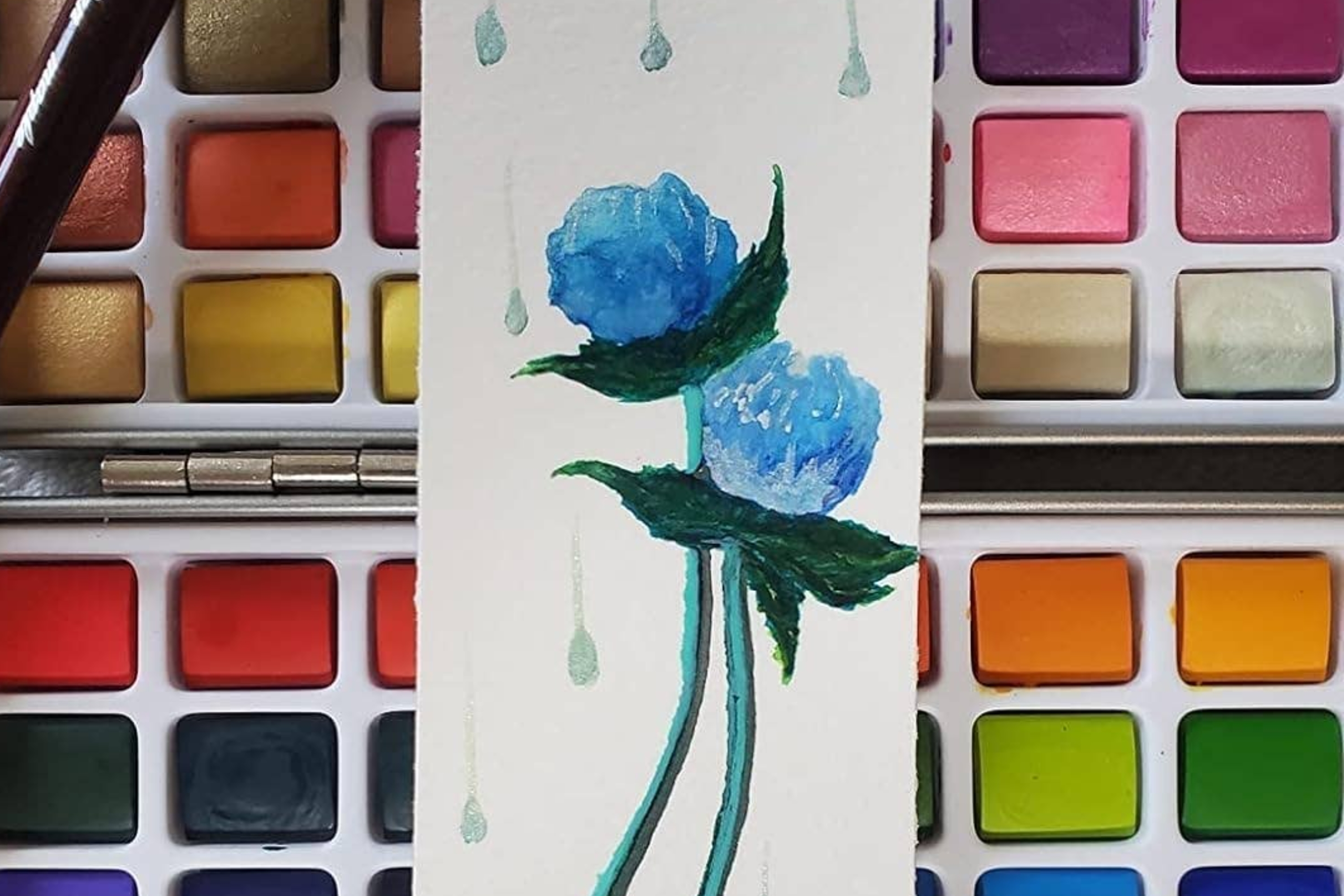 Watercolor painting of two blue flowers on a bookmark with a colorful paint palette in the background