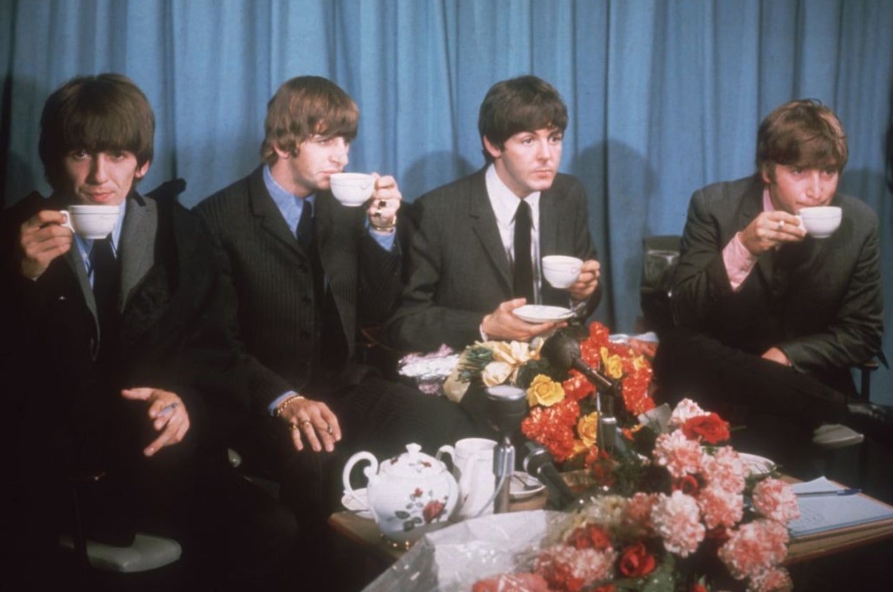 The Beatles in suits drink tea at a table with flowers and a teapot in a formal setting