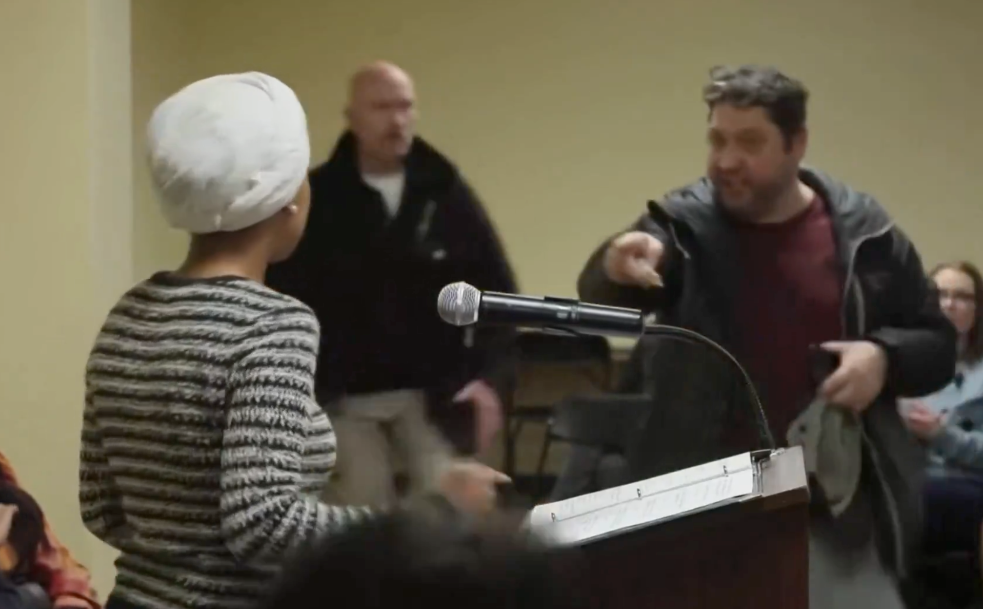 En person konfronterer en taler på et offentlig møte og gestikulerer med hånden. Høyttaleren, med tildekket hode, står bak et podium med mikrofon