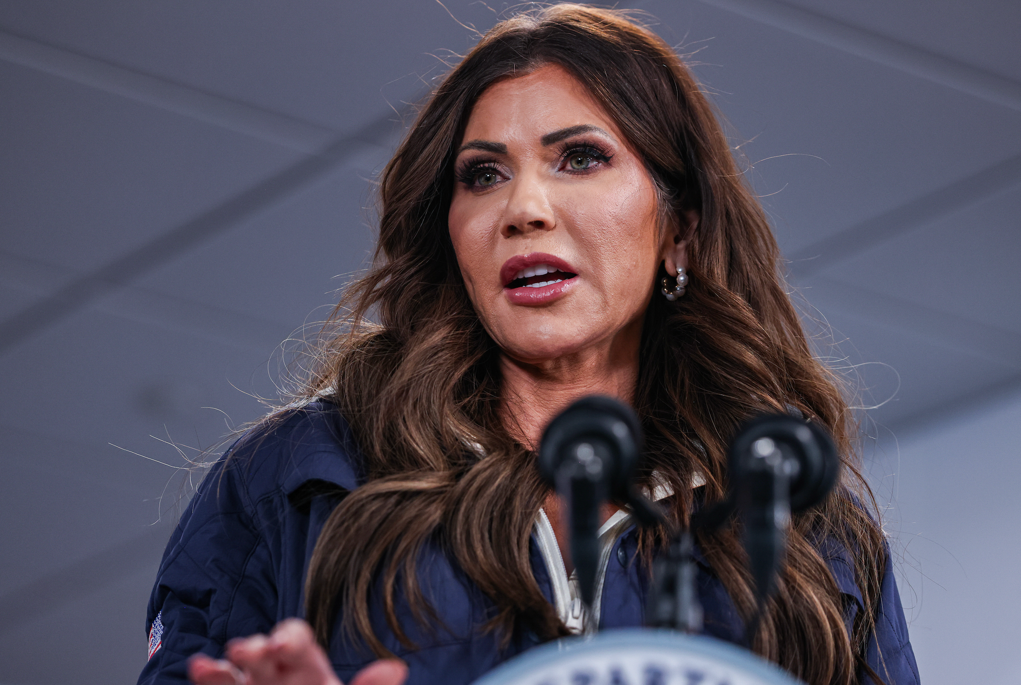 Person som snakker ved et podium med to mikrofoner, iført en formell jakke og øredobber, mot en innendørs bakgrunn