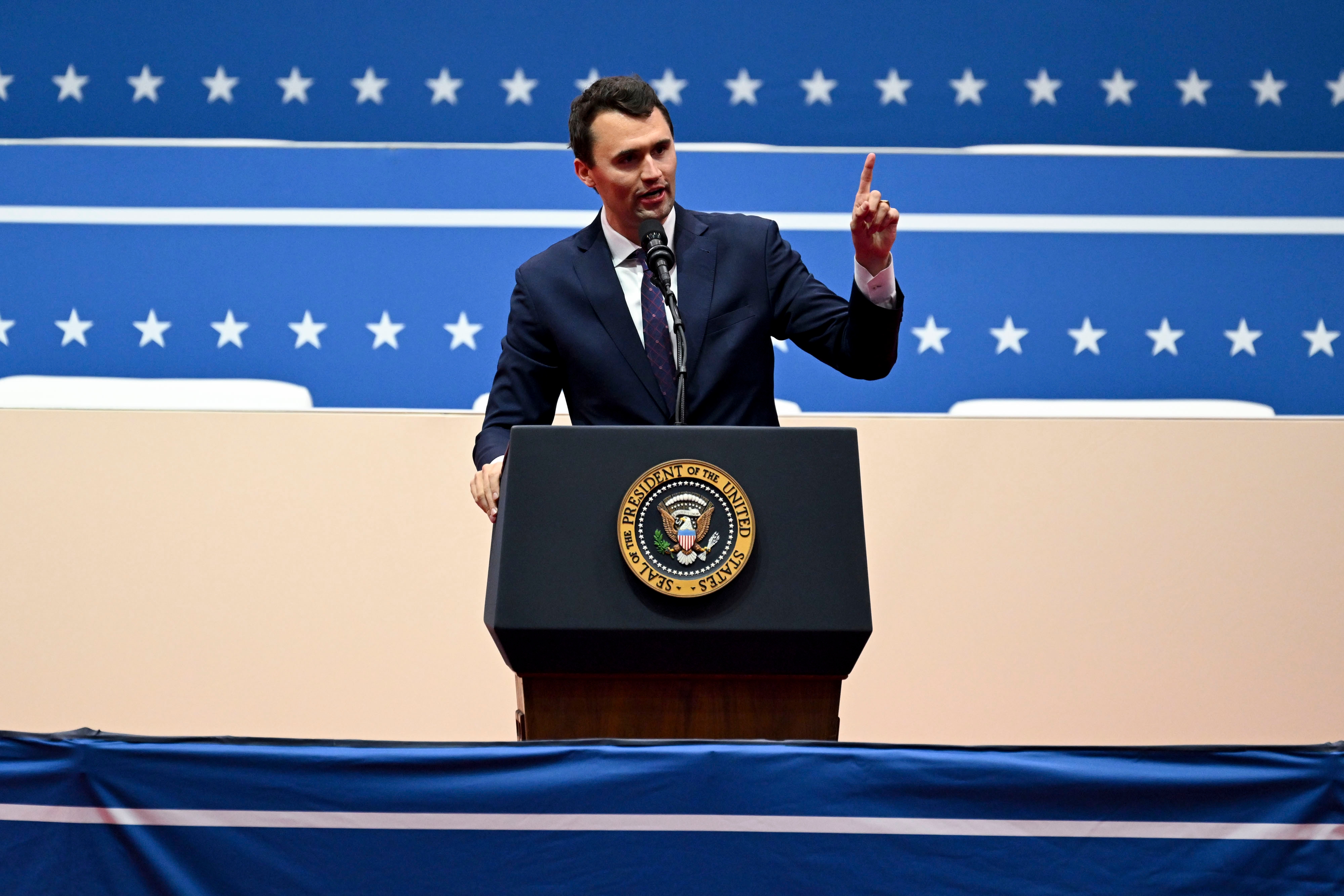 En mann i dress snakker lidenskapelig ved et podium med en presidentsegl under en formell begivenhet