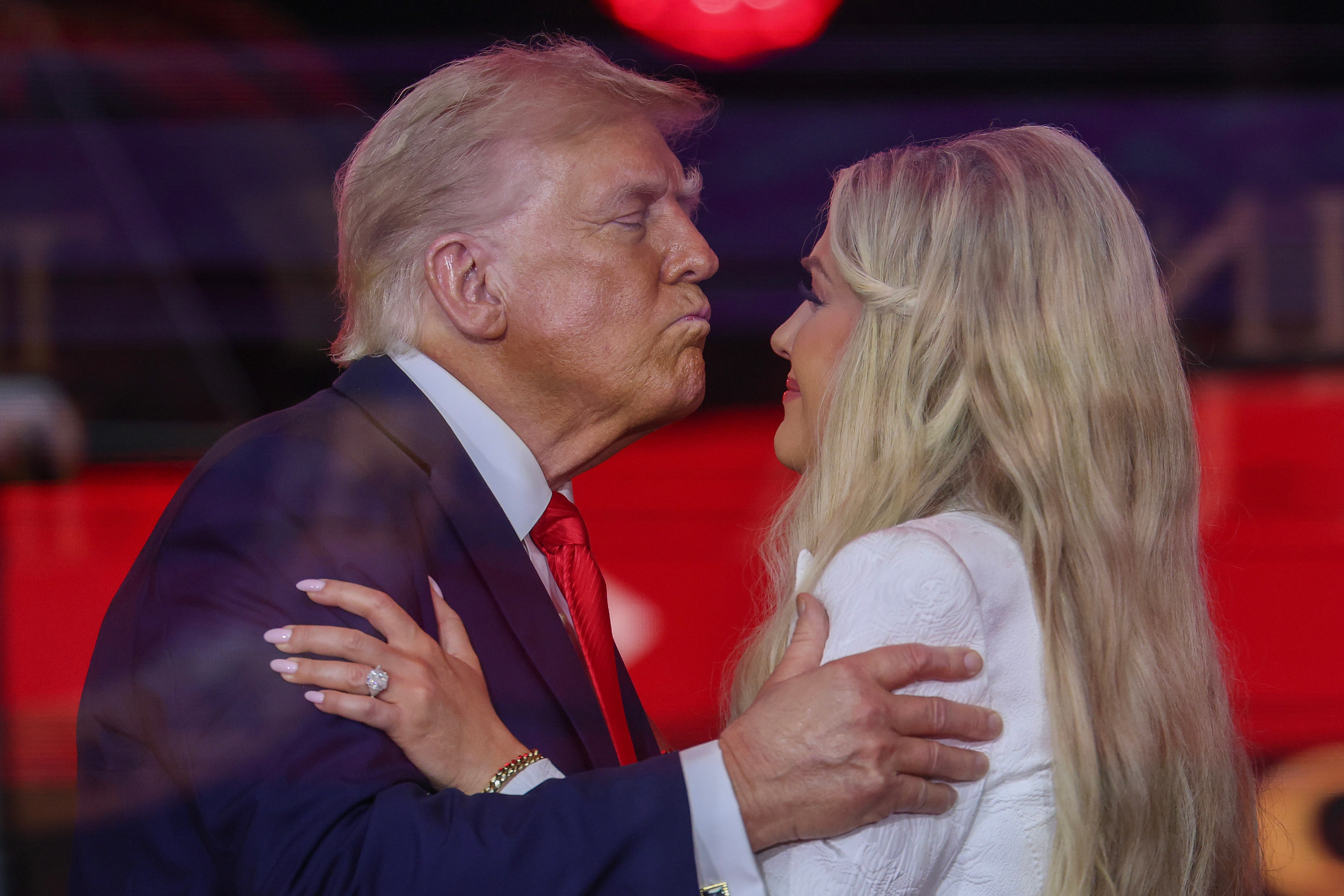 To personer på en scene er i ferd med å omfavne. Den ene har på seg dress, og den andre har hvitt antrekk. De er fremhevet mot et levende bakteppe