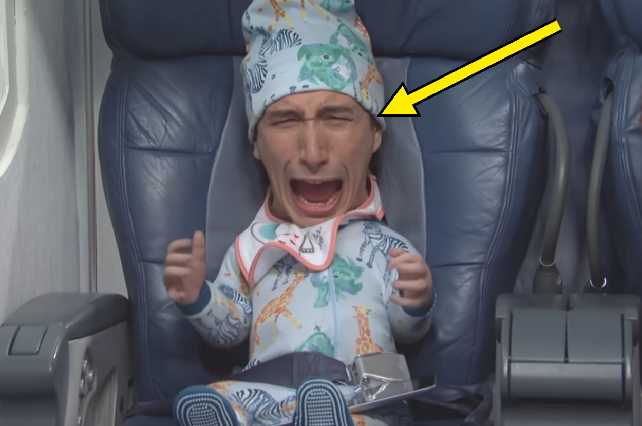 A passenger in a humorous baby costume cries while seated in an airplane, with a yellow arrow pointing toward their head
