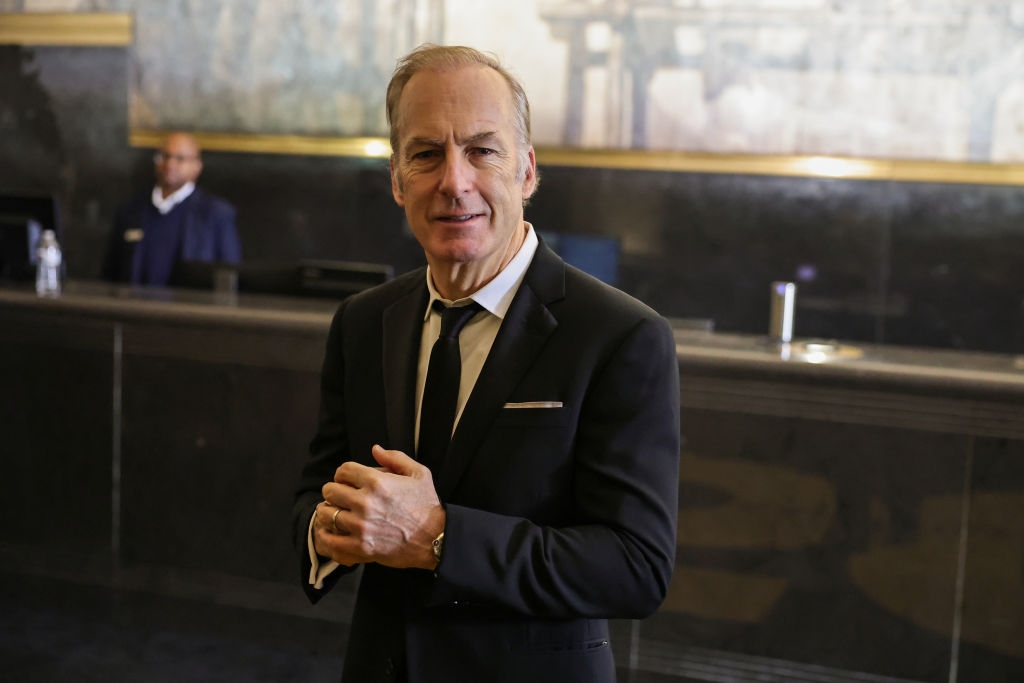 A man stands in a formal black suit in what appears to be a hotel lobby, with another person blurred in the background