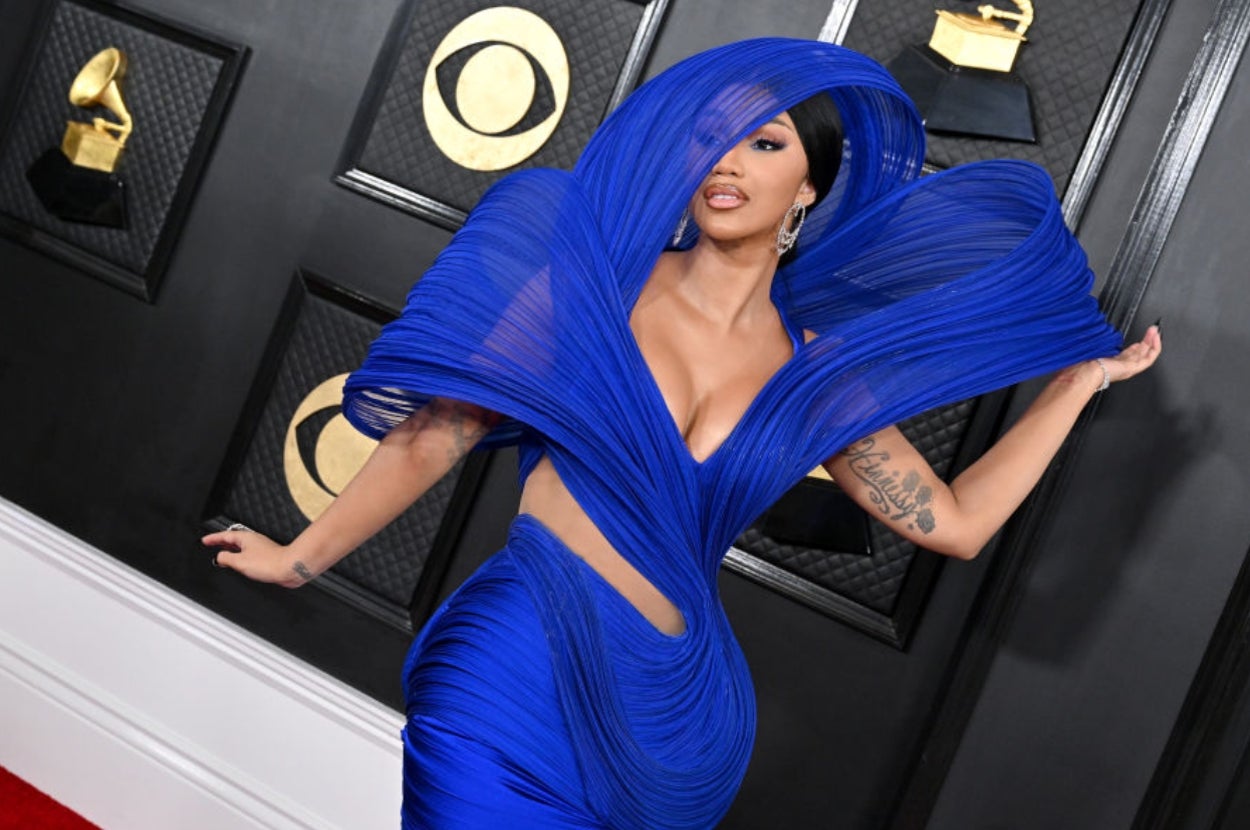 Person in a dramatic, sculptural gown poses on an awards red carpet surrounded by music-themed decor