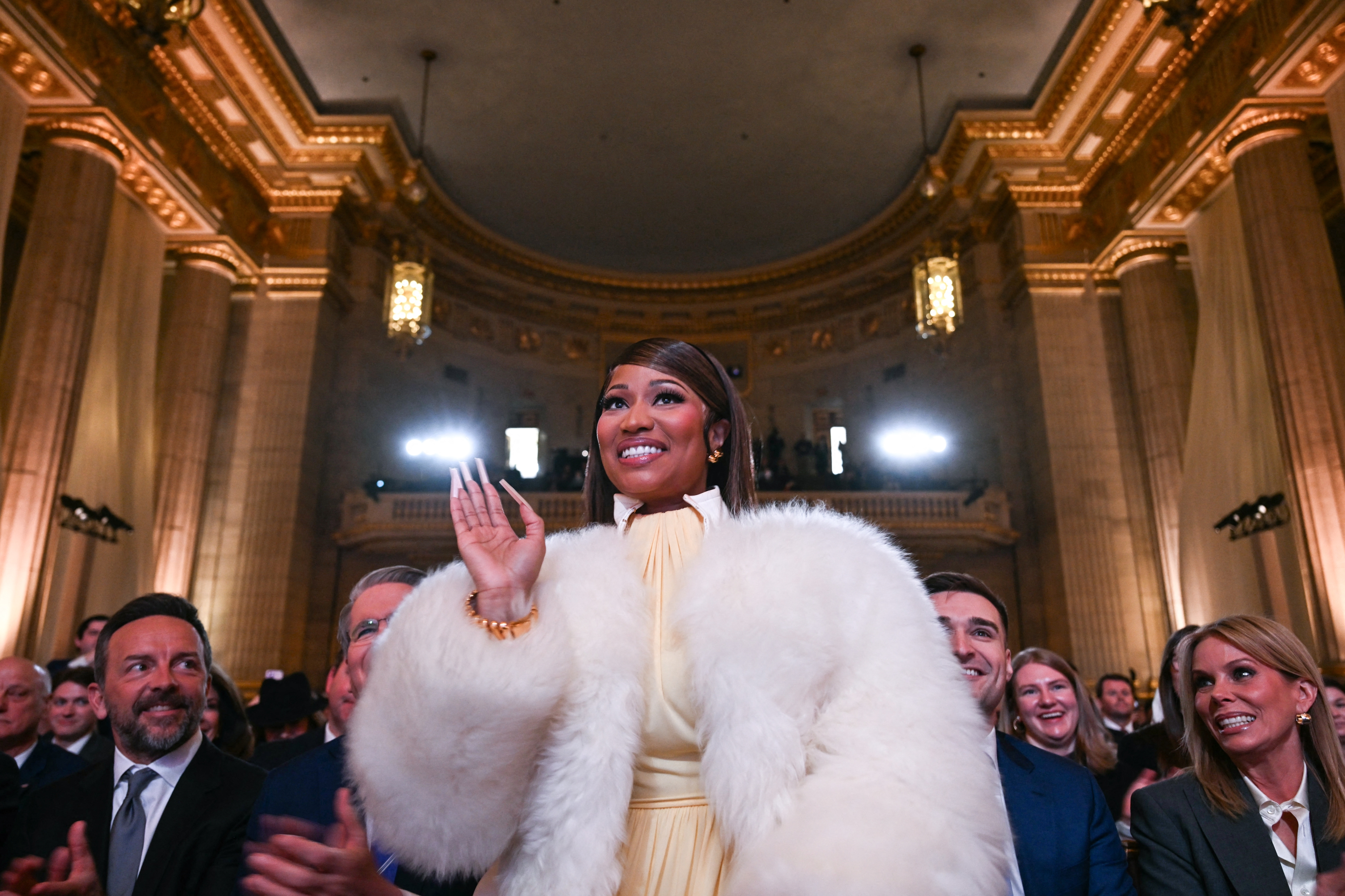 Person i en elegant kjole med pels vinker til publikum ved en formell begivenhet inne i en storslått, utsmykket hall. Publikum applauderer
