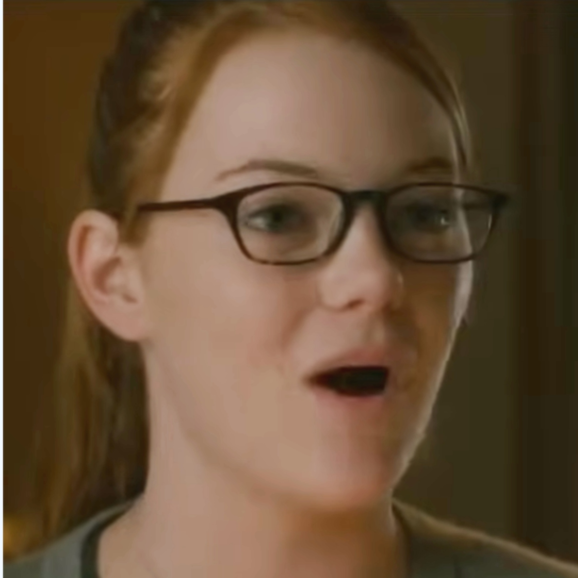 A person with glasses smiles and talks in a casual setting, wearing a black top and grey outerwear, with hair tied back