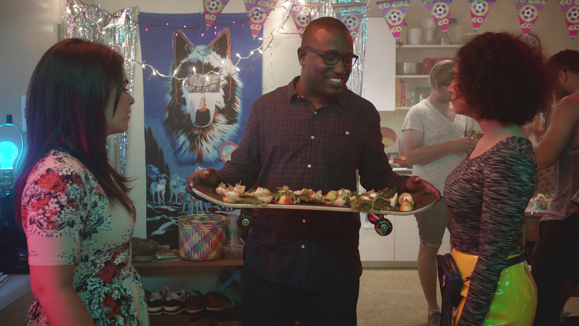 Person at a party holding a skateboard with snacks, talking to two others. A wolf poster and festive decorations are in the background