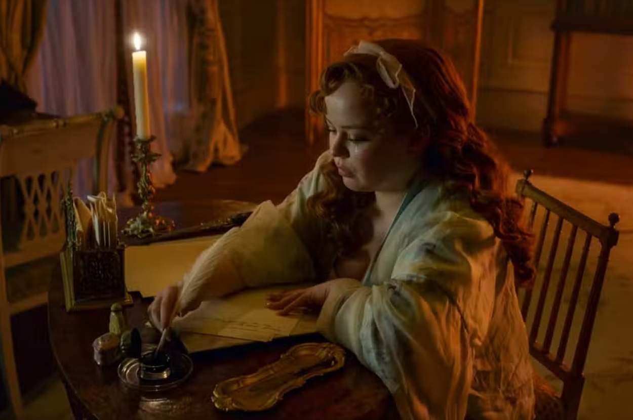 Penelope Bridgerton in period costume writing by candlelight at a desk with an ink quill, in a dimly lit room