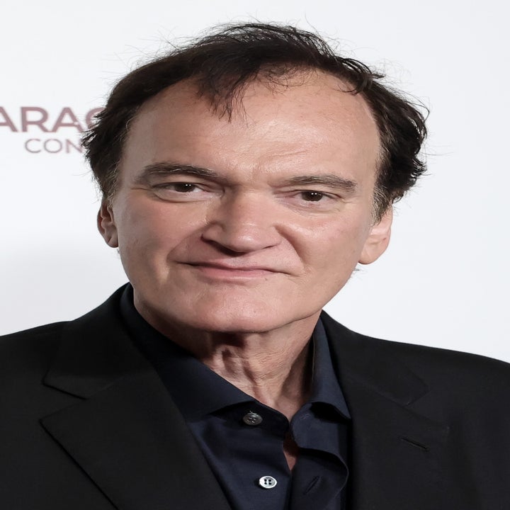 A person in a suit poses on a red carpet at an event, smiling with hands together