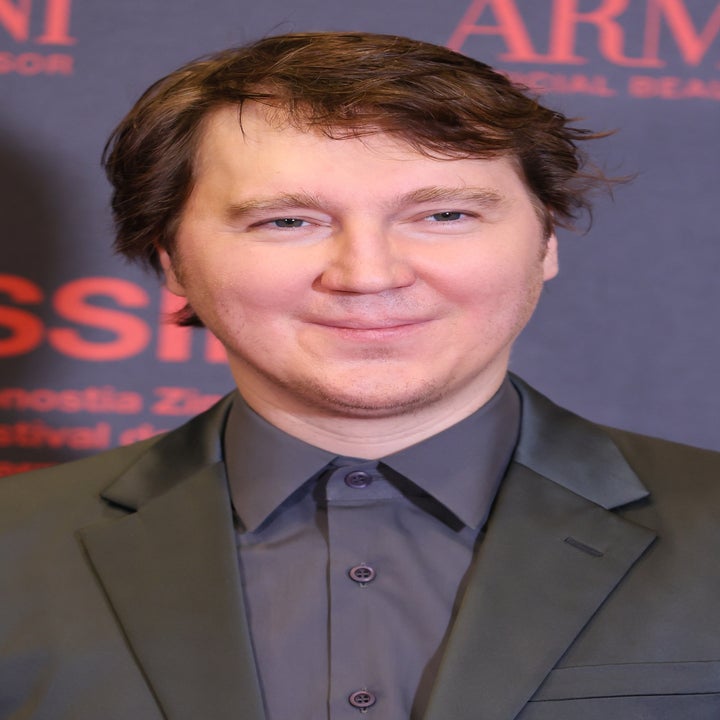 Person on red carpet wearing a dark suit with a subtle sheen, smiling against a backdrop with sponsor logos