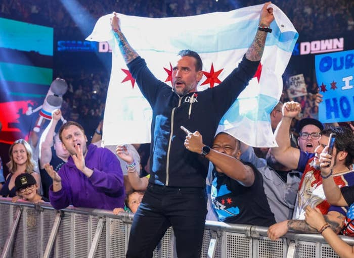 Wrestler holding a Chicago flag on a WWE stage, surrounded by excited fans with signs, celebrating energetically