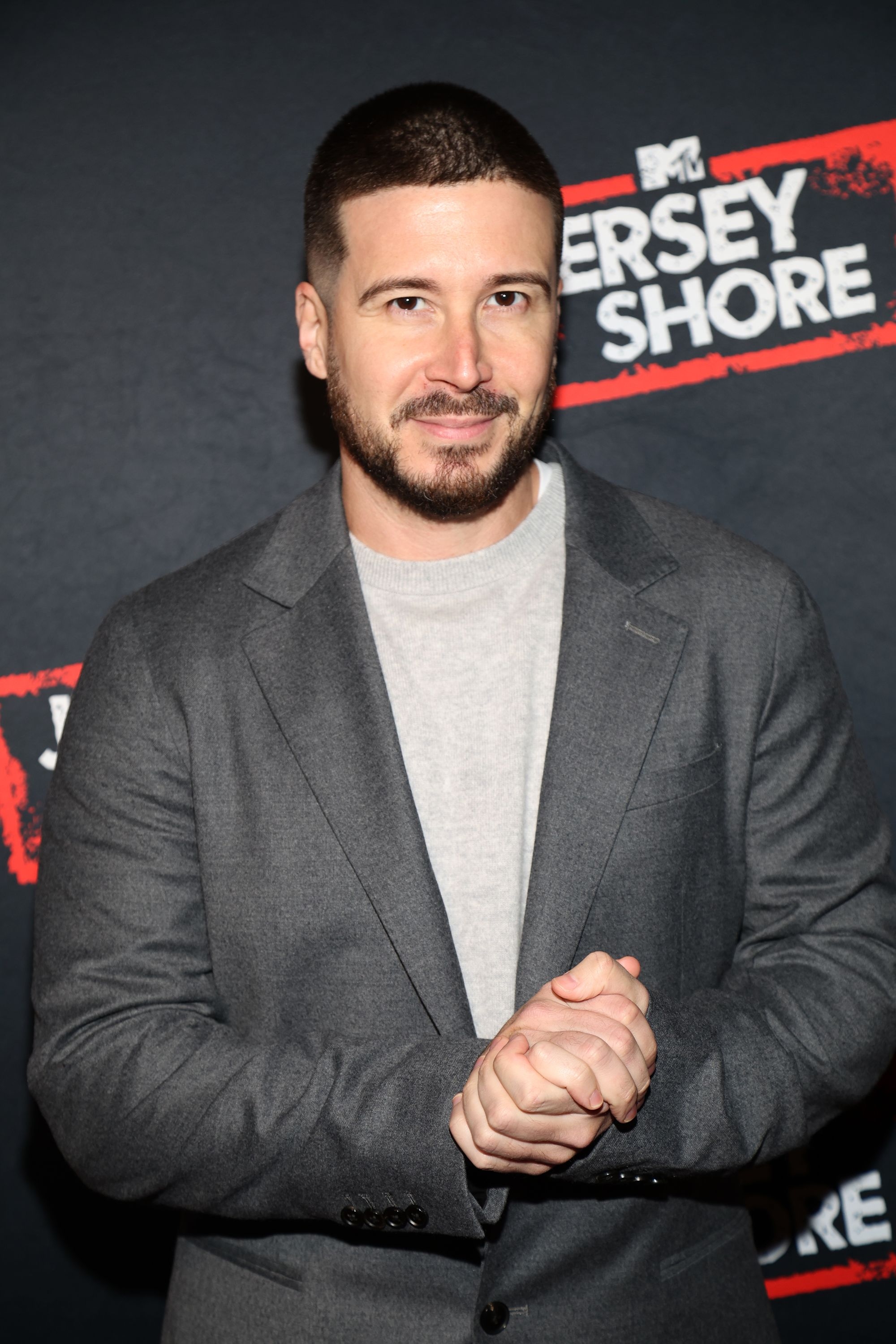 Vinny Guadagnino poses on a red carpet wearing a casual suit jacket over a light shirt with an MTV Jersey Shore backdrop