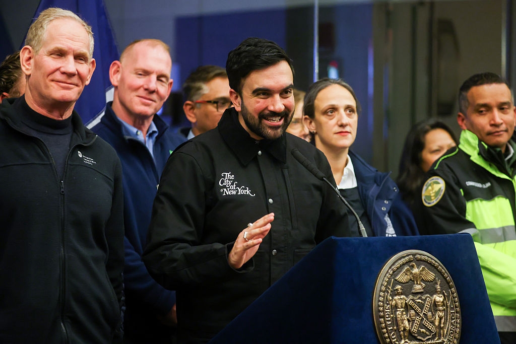 Gruppe embetsmenn på en pressekonferanse, sentertaler i jakke med «The City of New York»; emblem, smiler og gestikulerer mens de adresserer media