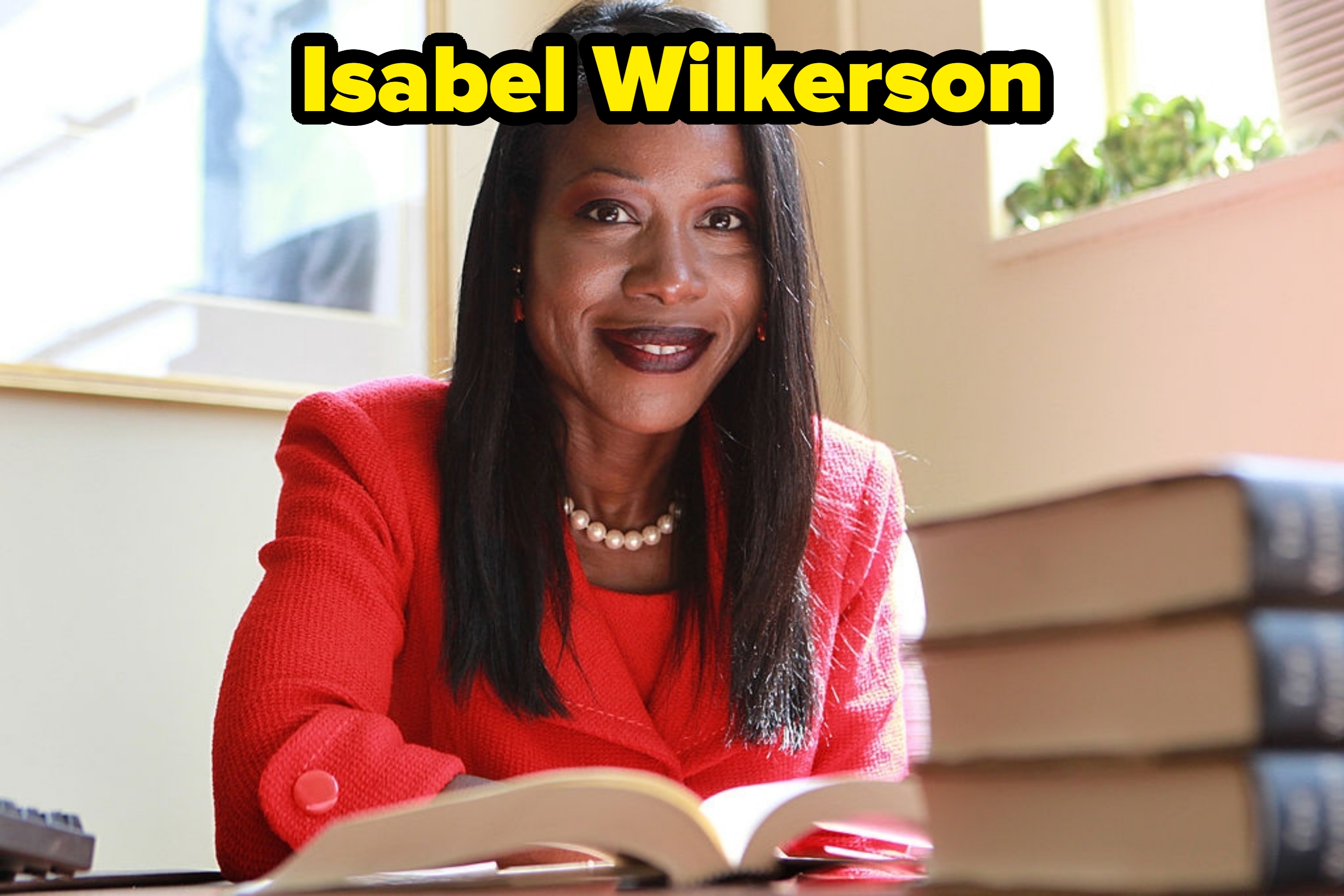 Person som sitter ved et bord med bøker, iført et formelt antrekk og perlekjede, smiler