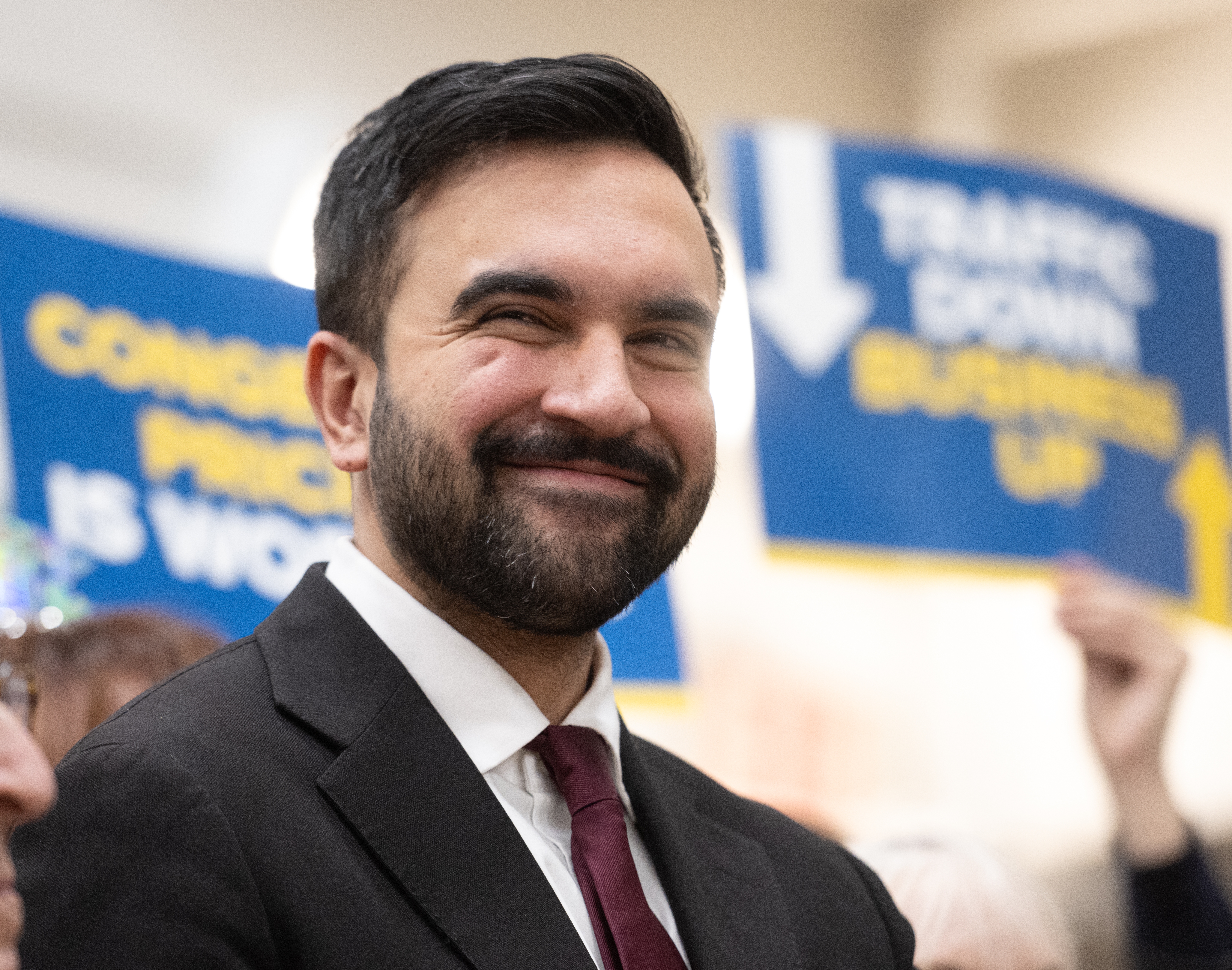 Mann i dress og slips smiler på et arrangement. Uskarpe bakgrunnsskilt antyder en forretningsmessig eller politisk kontekst