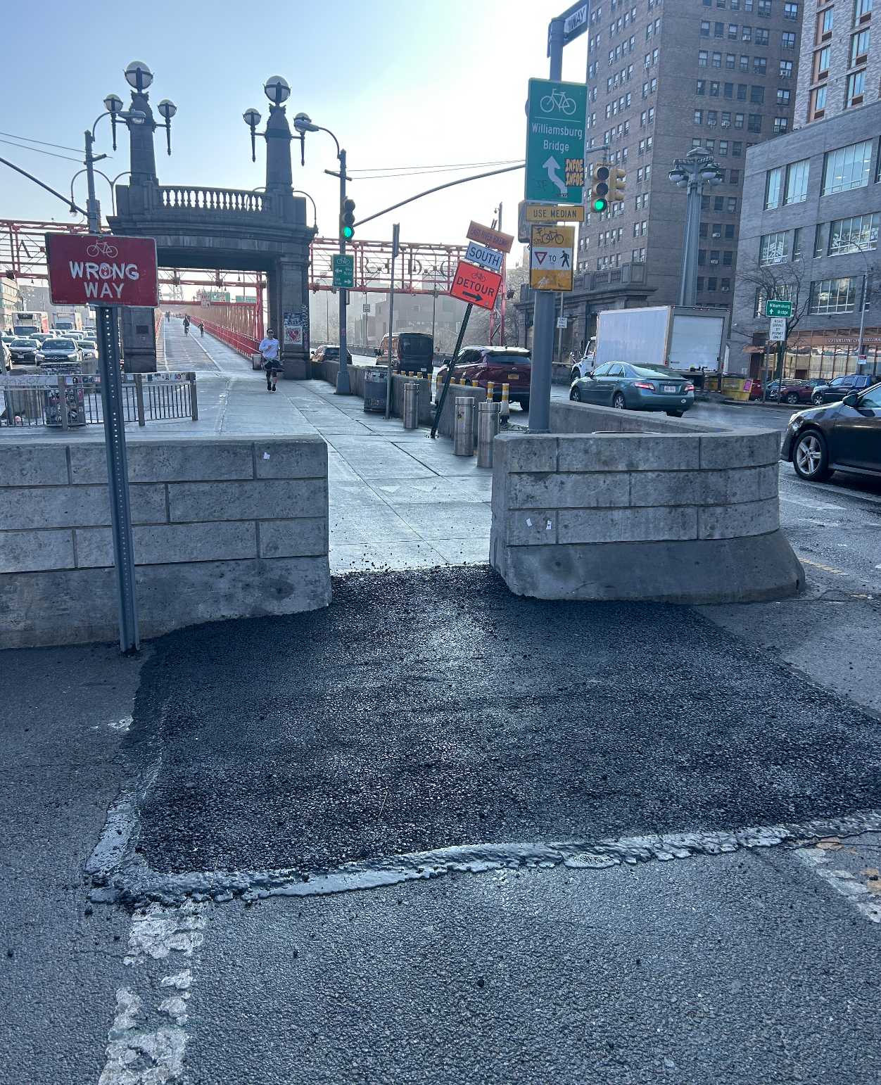 Betongbarrierer blokkerer et kjørefelt nær en bybro, med synlige trafikkskilt og en gate som strekker seg i det fjerne på en solrik dag