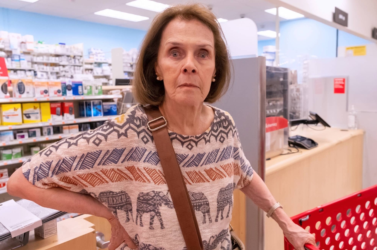 A person stands in a store aisle with a serious expression, holding a shopping cart. Shelves with products line the background