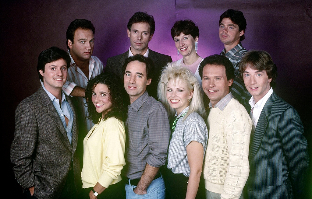 Group of smiling people in 1980s fashion, including blazers, sweaters, and shirts