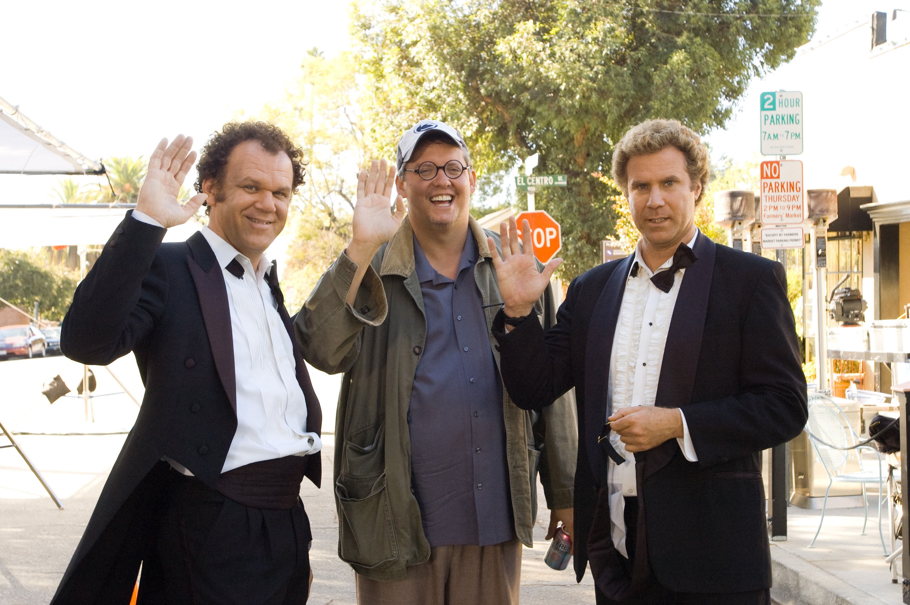 Three men, two in tuxedos and one in casual attire with a cap, smile and wave at the camera outdoors on a street