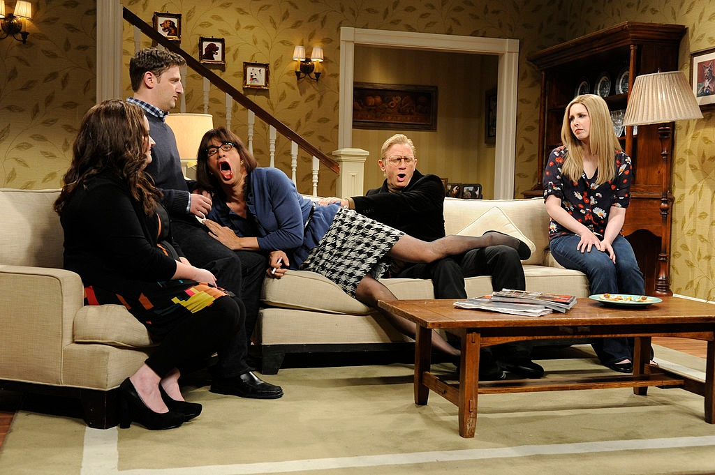 A comedic scene with four people on a couch in a sitcom set. One person is sprawled dramatically, entertaining the group; others watch with mixed reactions