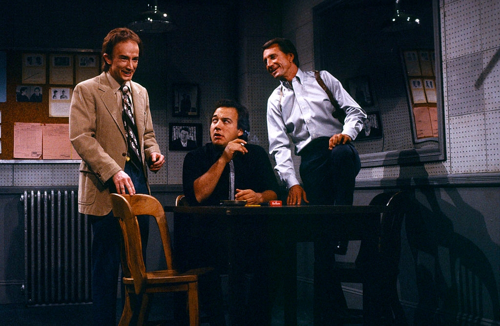 Three men in a dimly lit room, dressed in vintage business attire, stand and sit around a wooden table, engaged in conversation