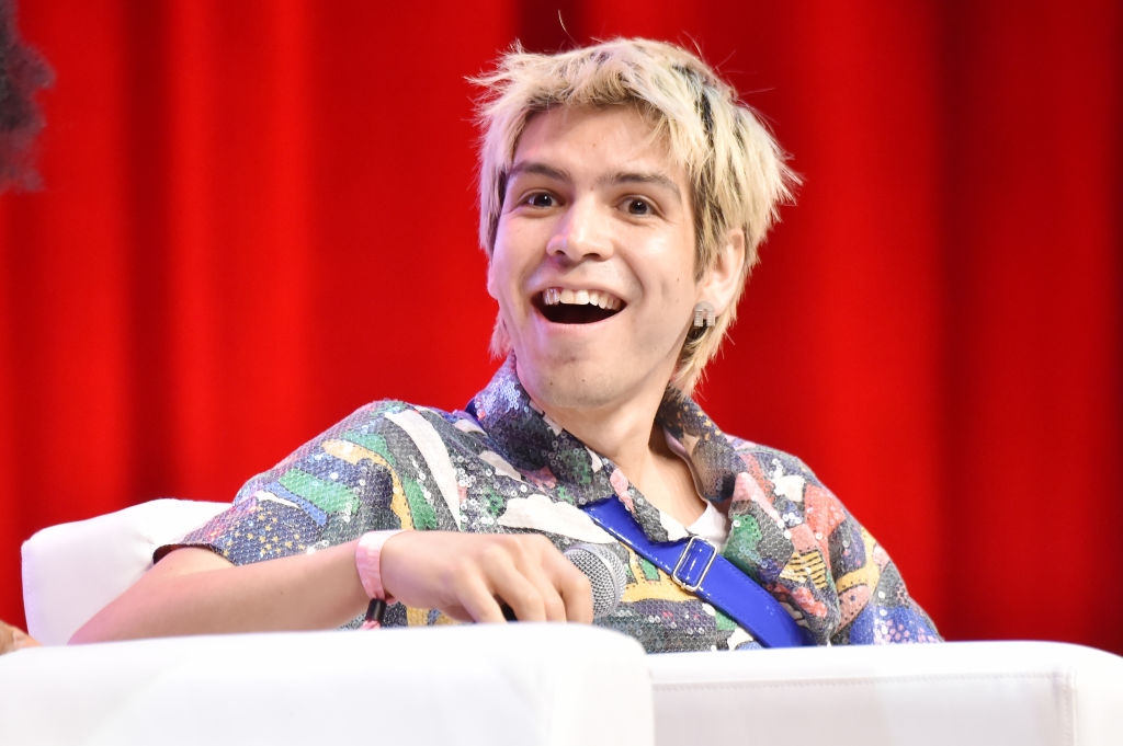 Person with a patterned shirt smiling while sitting on a panel with a red backdrop in the background