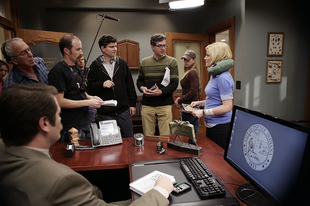 Behind-the-scenes photo of a TV show production. Cast and crew in discussion around a desk with scripts and equipment visible