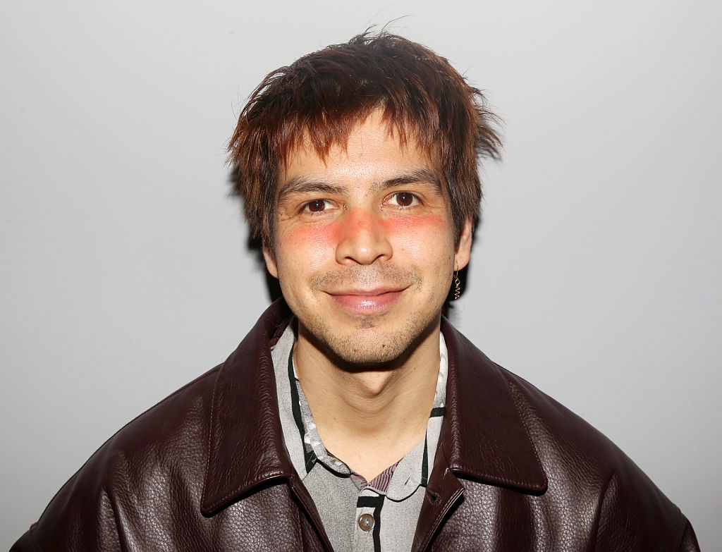 Person with a subtle smile wearing a leather jacket over a collared shirt