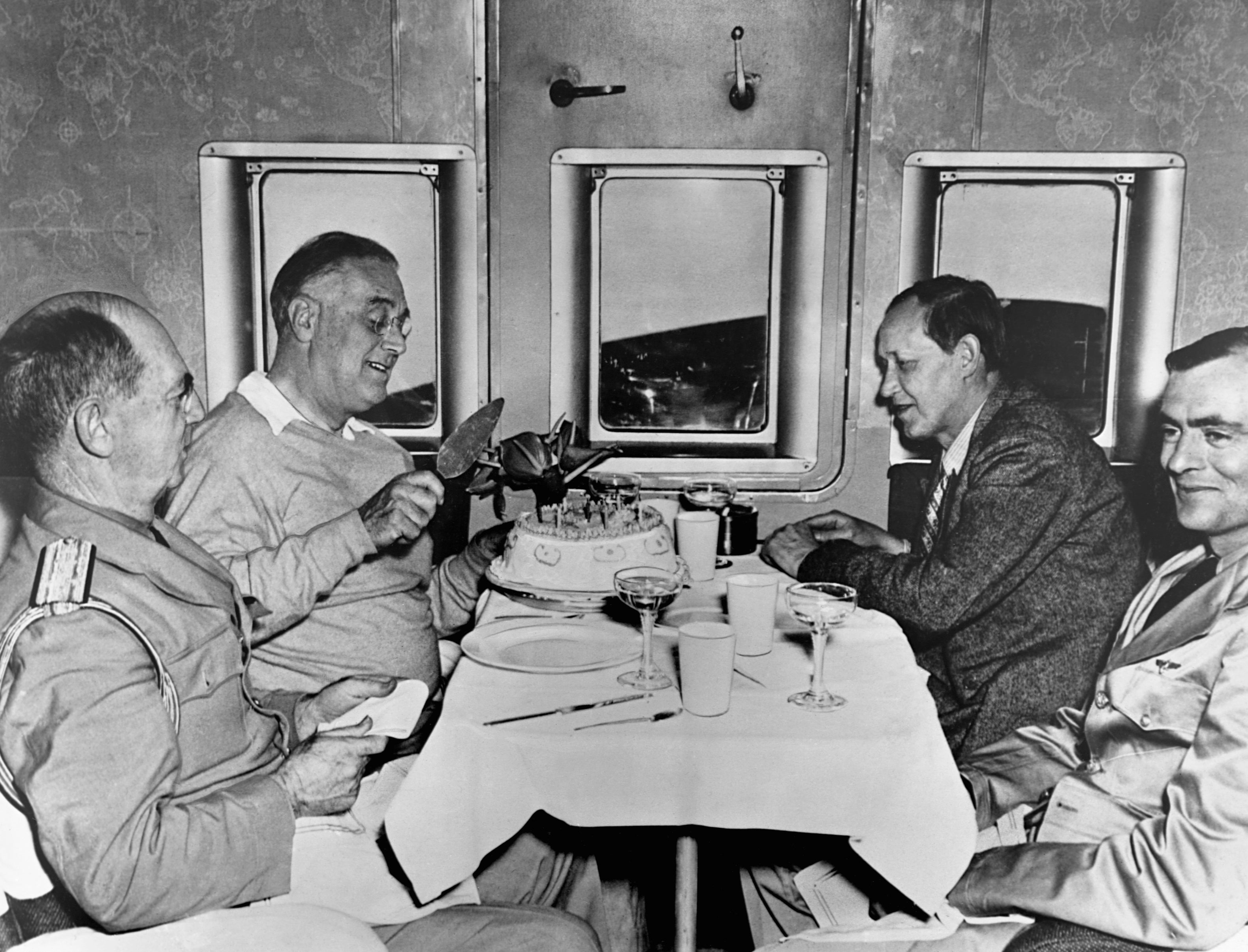 Four men successful  subject   attire dine astatine  a array  successful  an aircraft. One antheral   gestures towards a barroom   successful  the center