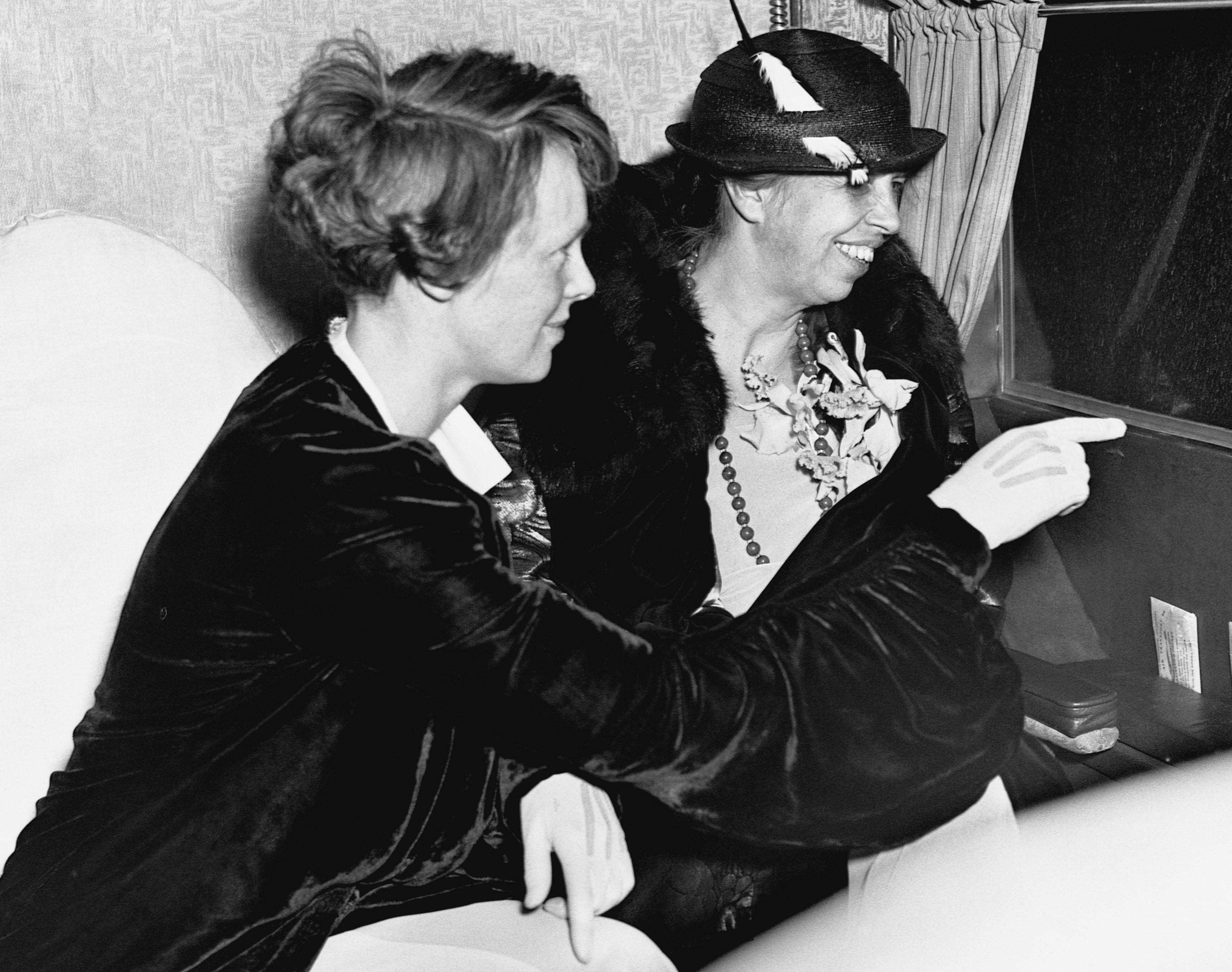 Two women from the 1930s, sitting and smiling. One points outward. Both deterioration  stylish hats and coats, indicating a ceremonial  oregon  question   setting
