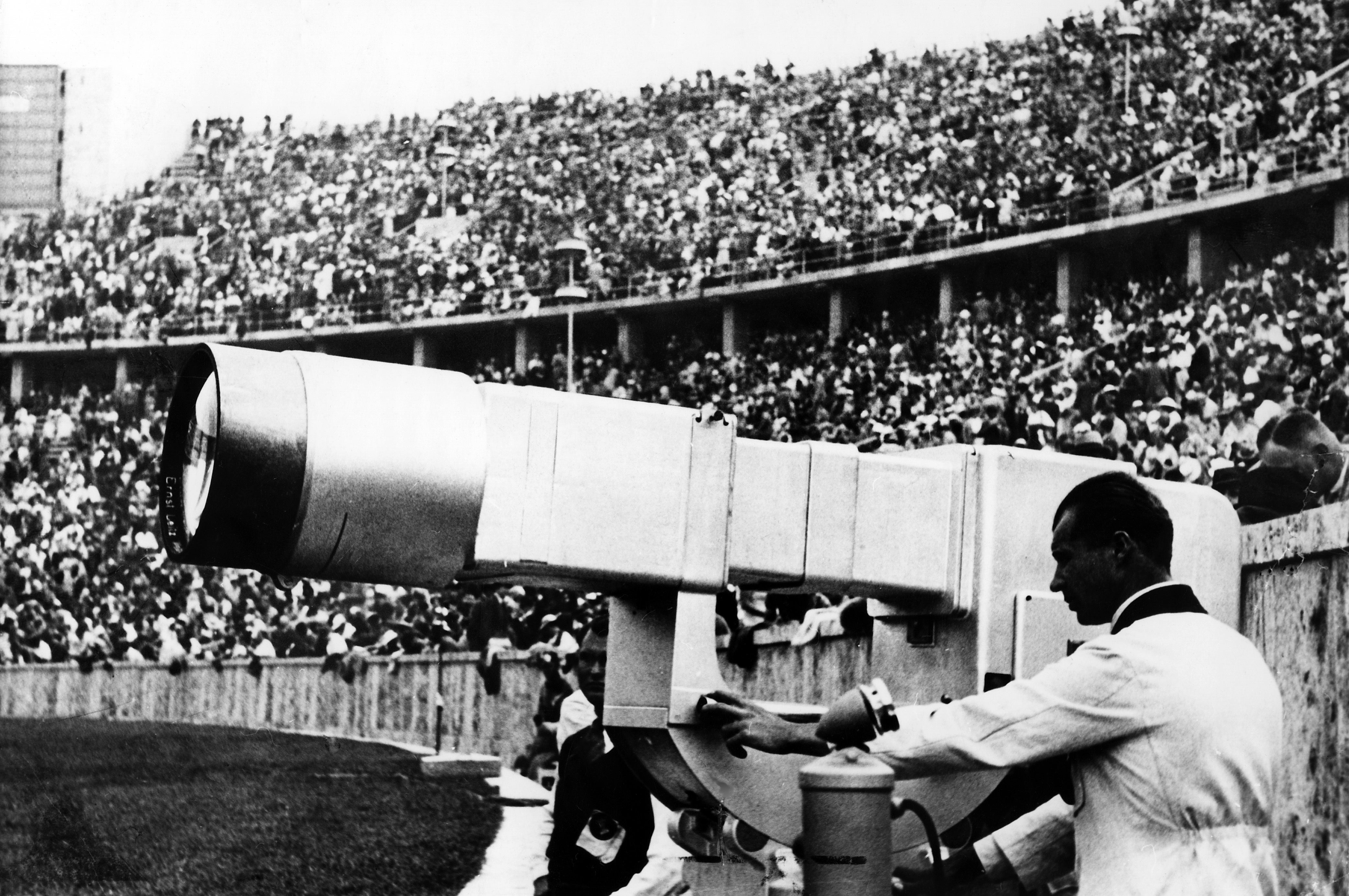A ample  assemblage  watches arsenic  a antheral   operates an aboriginal  tv  camera astatine  a stadium event, showcasing 20th-century broadcasting technology