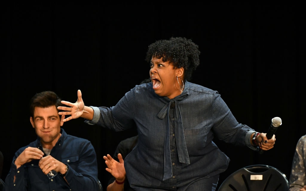 Woman performing energetically on stage, holding a microphone, wearing a denim outfit. Audience member sits behind her, reacting with amusement
