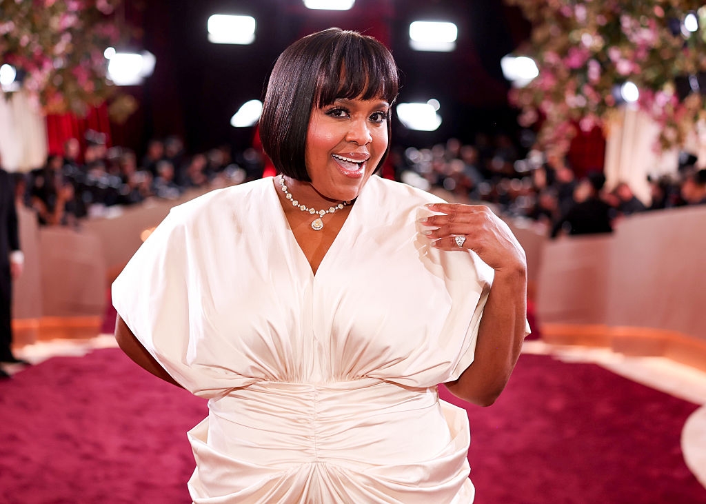 Person in elegant gown with short, sleek bob haircut on a red carpet. They're smiling, posing confidently with a hand on hip and visible ring