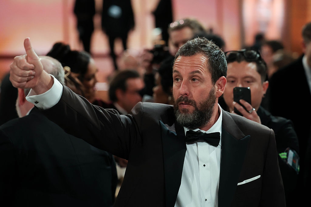 Man in a tuxedo gives a thumbs up on a red carpet, surrounded by people and photographers
