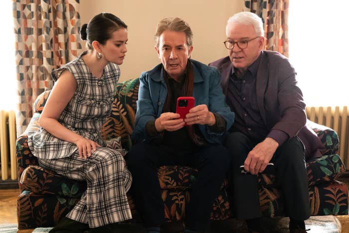 Three people sit on a couch, looking at a phone together. The person on the left wears a patterned outfit, the middle person holds the phone, and the right person wears a blazer