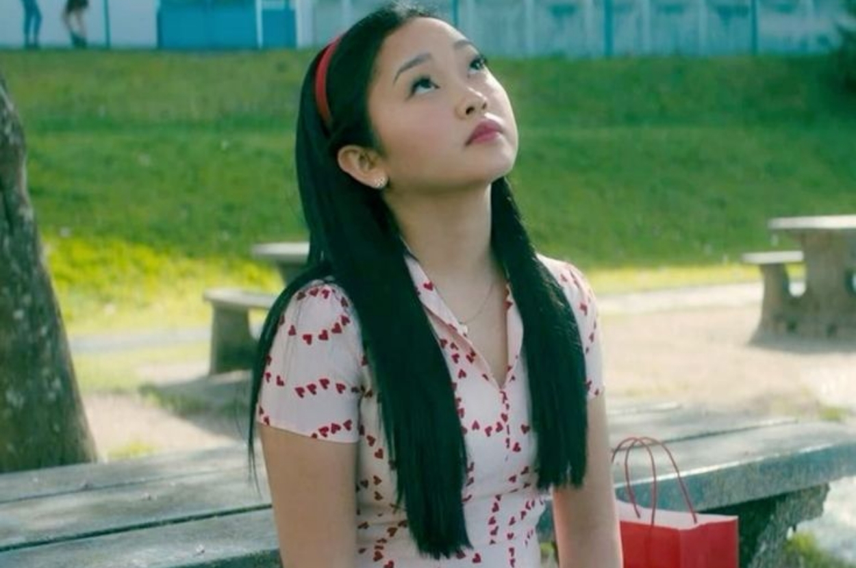 A person with long hair sits on a bench outdoors, looking up contemplatively. They wear a heart-patterned dress and a headband