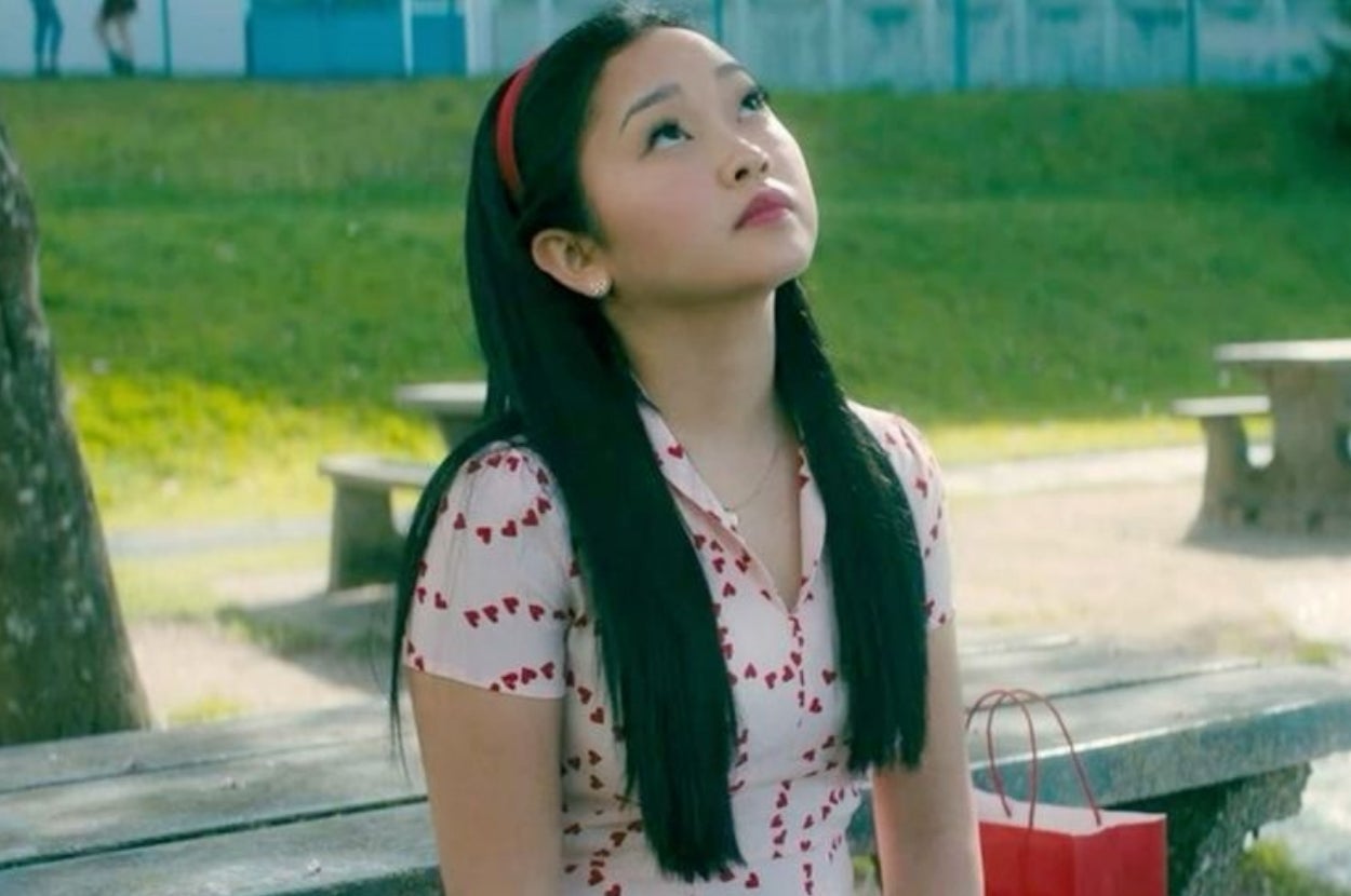 A person with long hair sits on a bench outdoors, looking up contemplatively. They wear a heart-patterned dress and a headband