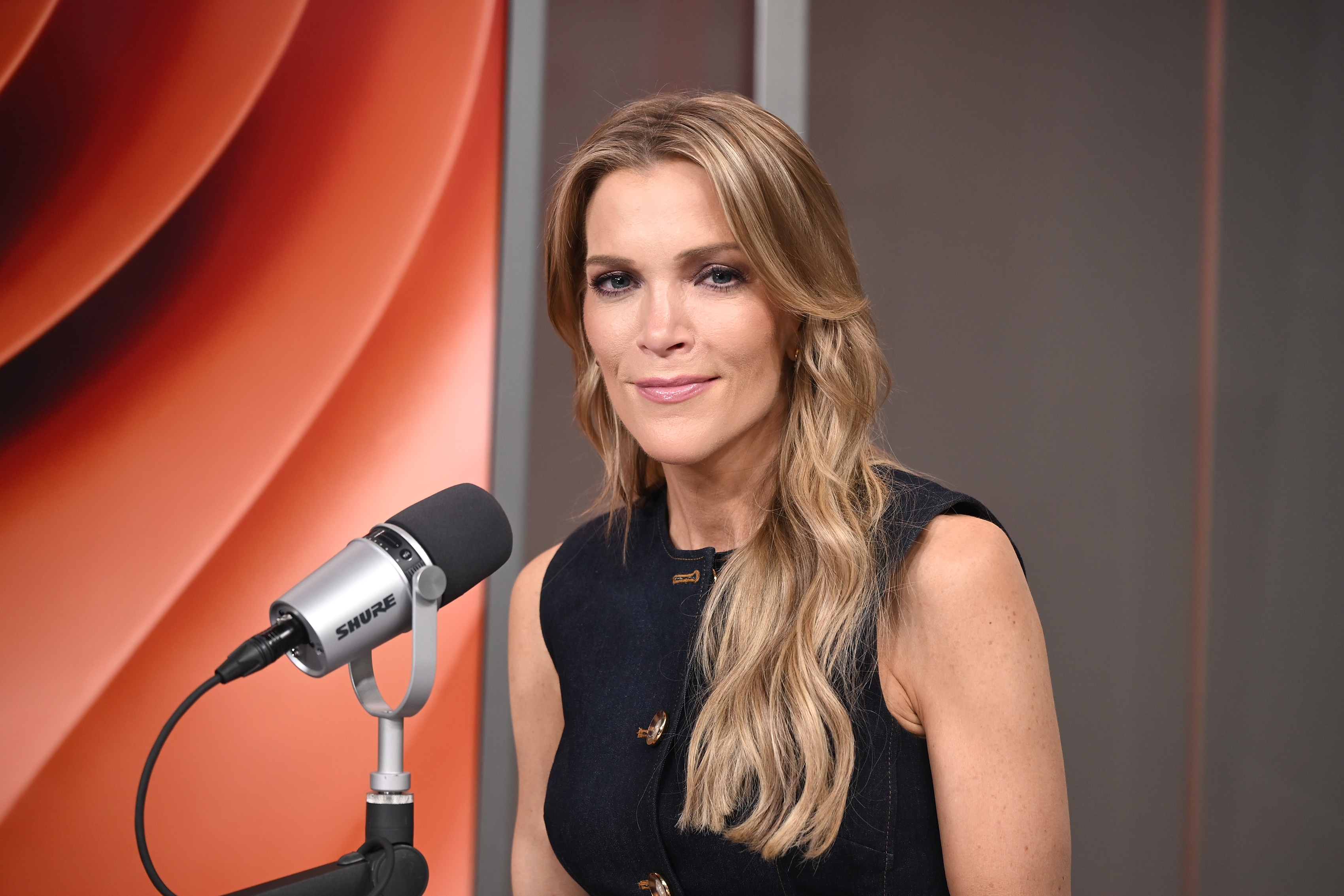 En person med langt hår, i et ermeløst svart antrekk, sitter ved en mikrofon og foreslår en podcast eller intervjuinnstilling