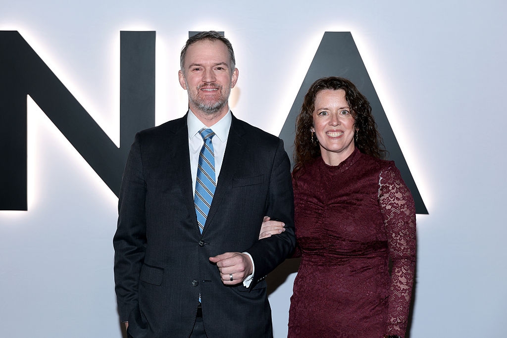 Mann i dress med stripet slips og kvinne i blondekjole poserer sammen på et arrangement, smilende