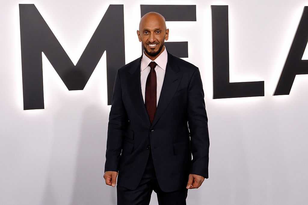 En person i en formell mørk dress og slips på en rød løper, smilende for bilder foran et bakteppe med store bokstaver