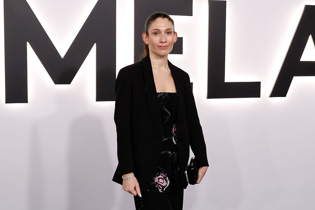 Person i stilig svart antrekk med mønstret topp, stående foran et bakteppe med store bokstaver