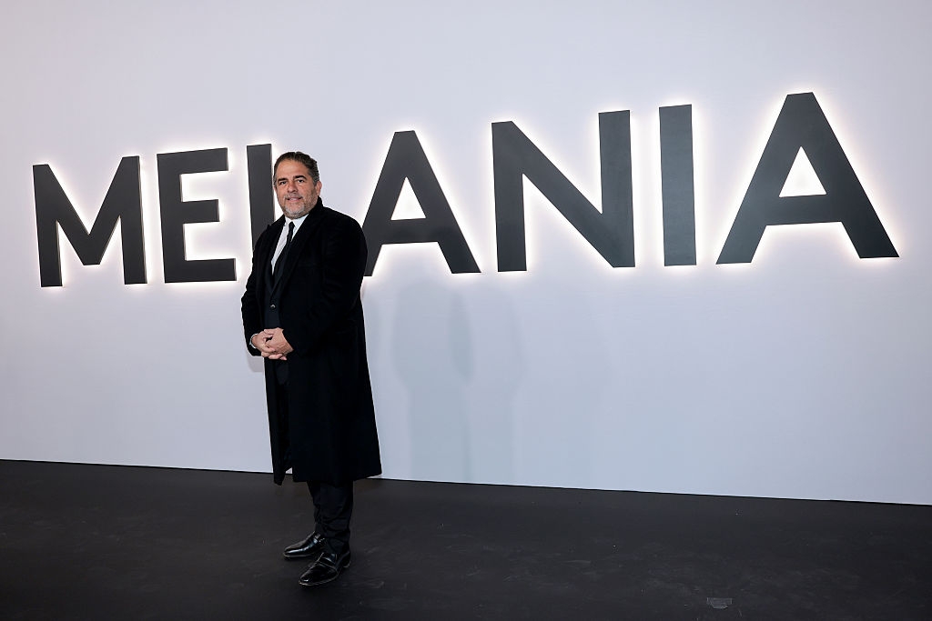 En mann i formell dress og frakk står foran en stor "MELANIA" signere på et arrangement