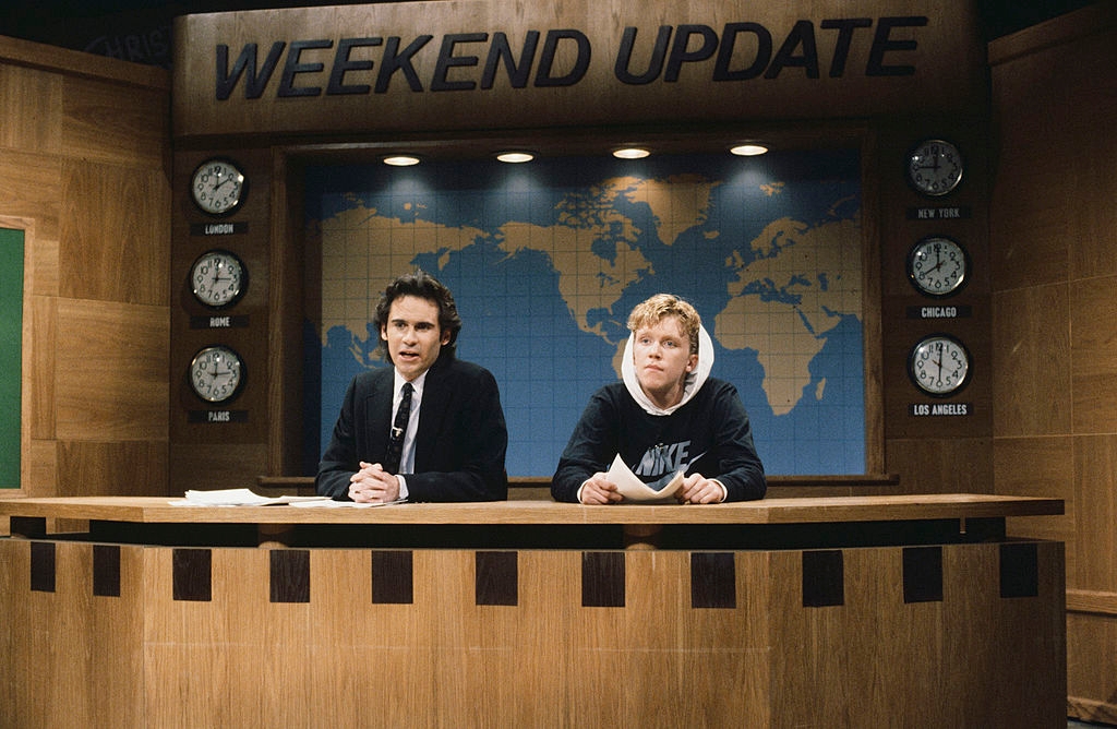 Two people sitting at a "Weekend Update" news desk. One in a suit, the other in a Nike hoodie, with a world map and clocks in the background
