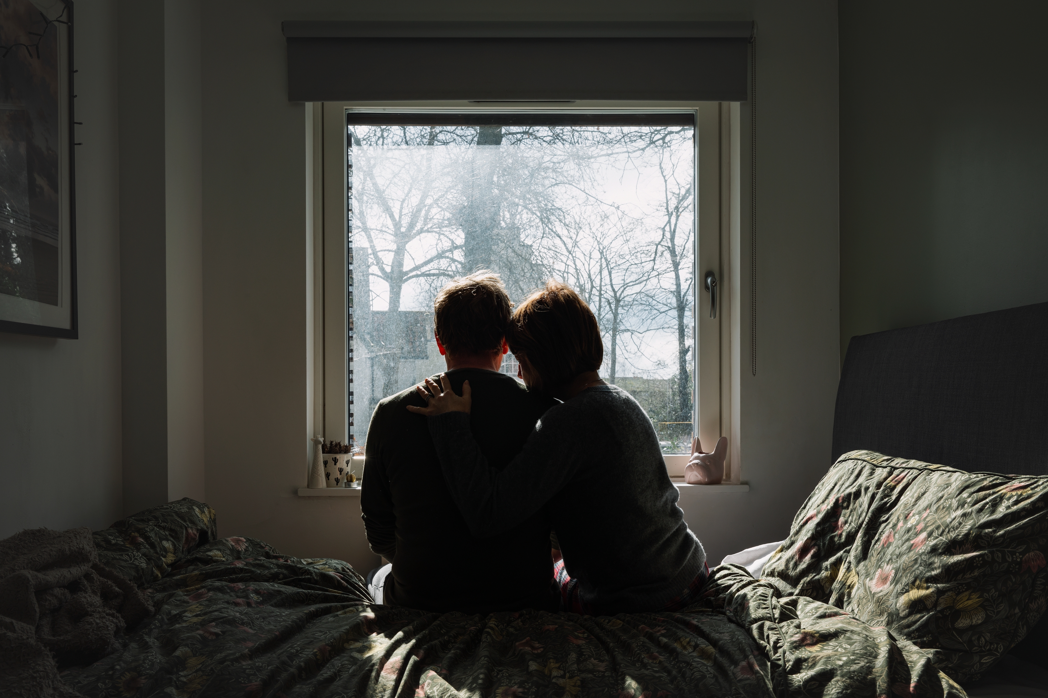 Two people sit closely together on a bed, facing a window with trees visible outside, creating an intimate and serene atmosphere