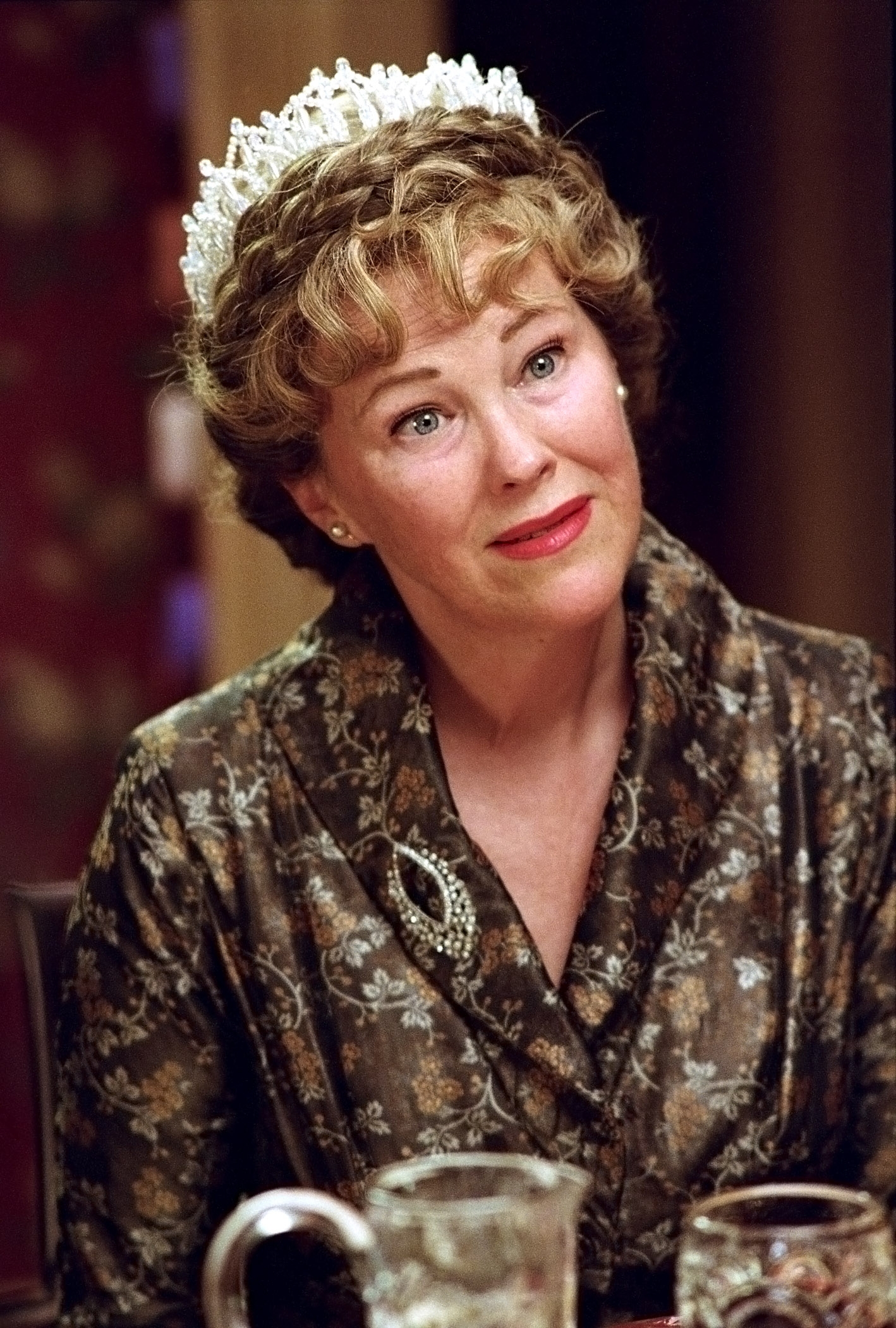 Person wearing a patterned coat with a brooch and a pearl tiara, sitting at a table, looking upward with a thoughtful expression