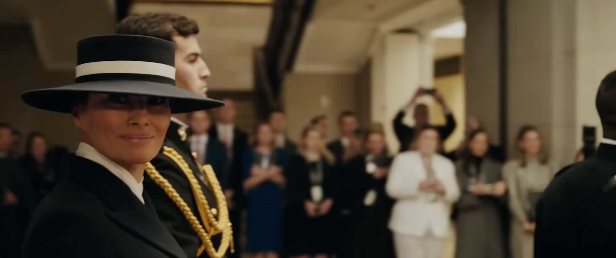 Person iført en bredbremmet hatt og skreddersydd dress på en formell begivenhet, omgitt av et publikum