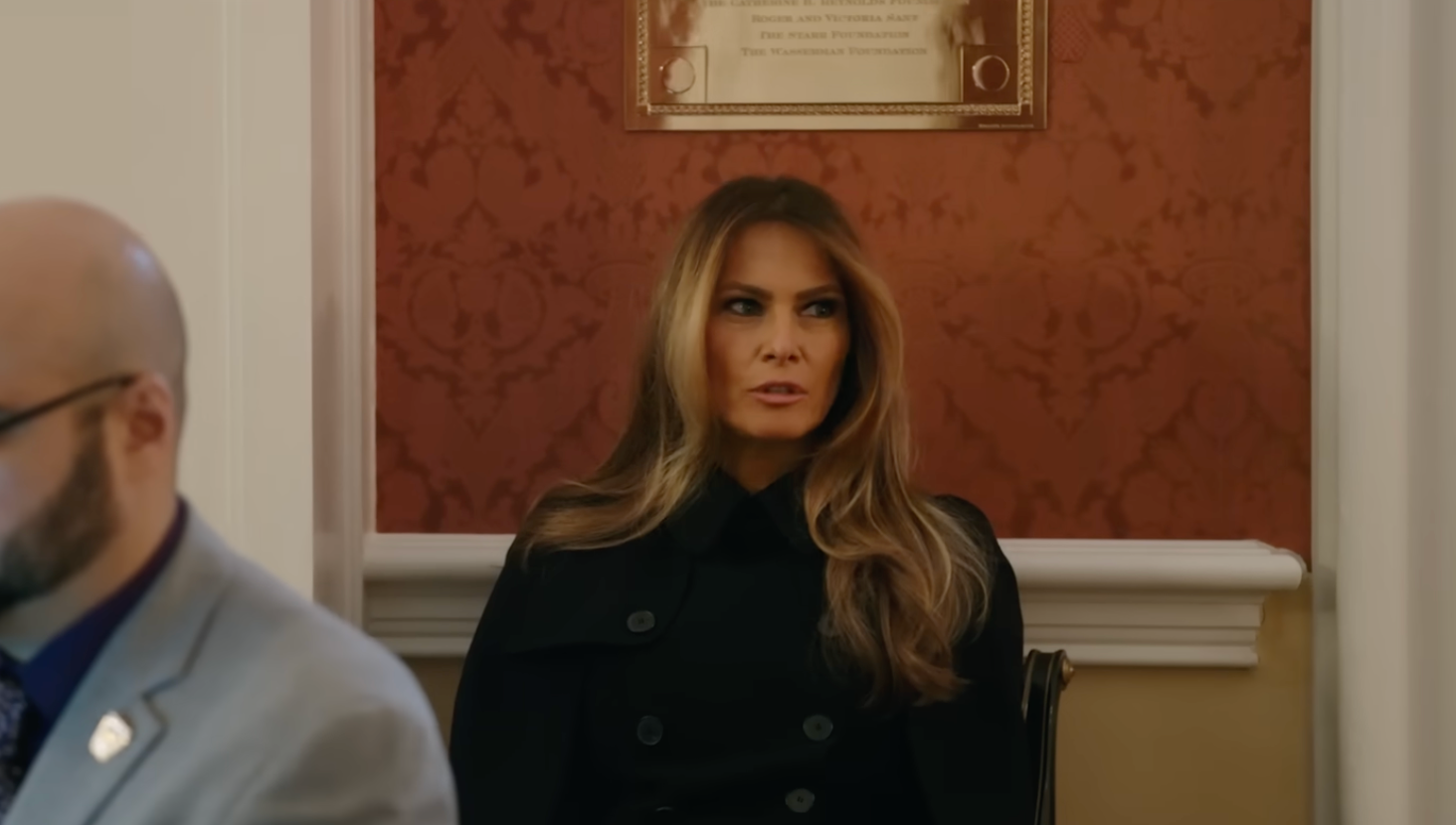 En kvinne i svart frakk sitter i en formell setting. En annen person i dress er delvis synlig i forgrunnen