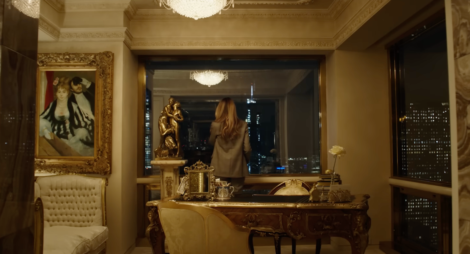 En person står i et overdådig rom og ser ut på en bysilhuett gjennom et stort vindu. Det er et gullinnrammet maleri og utsmykkede møbler